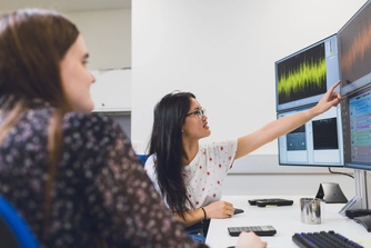 Two female students in front of computers displaying graphs and data